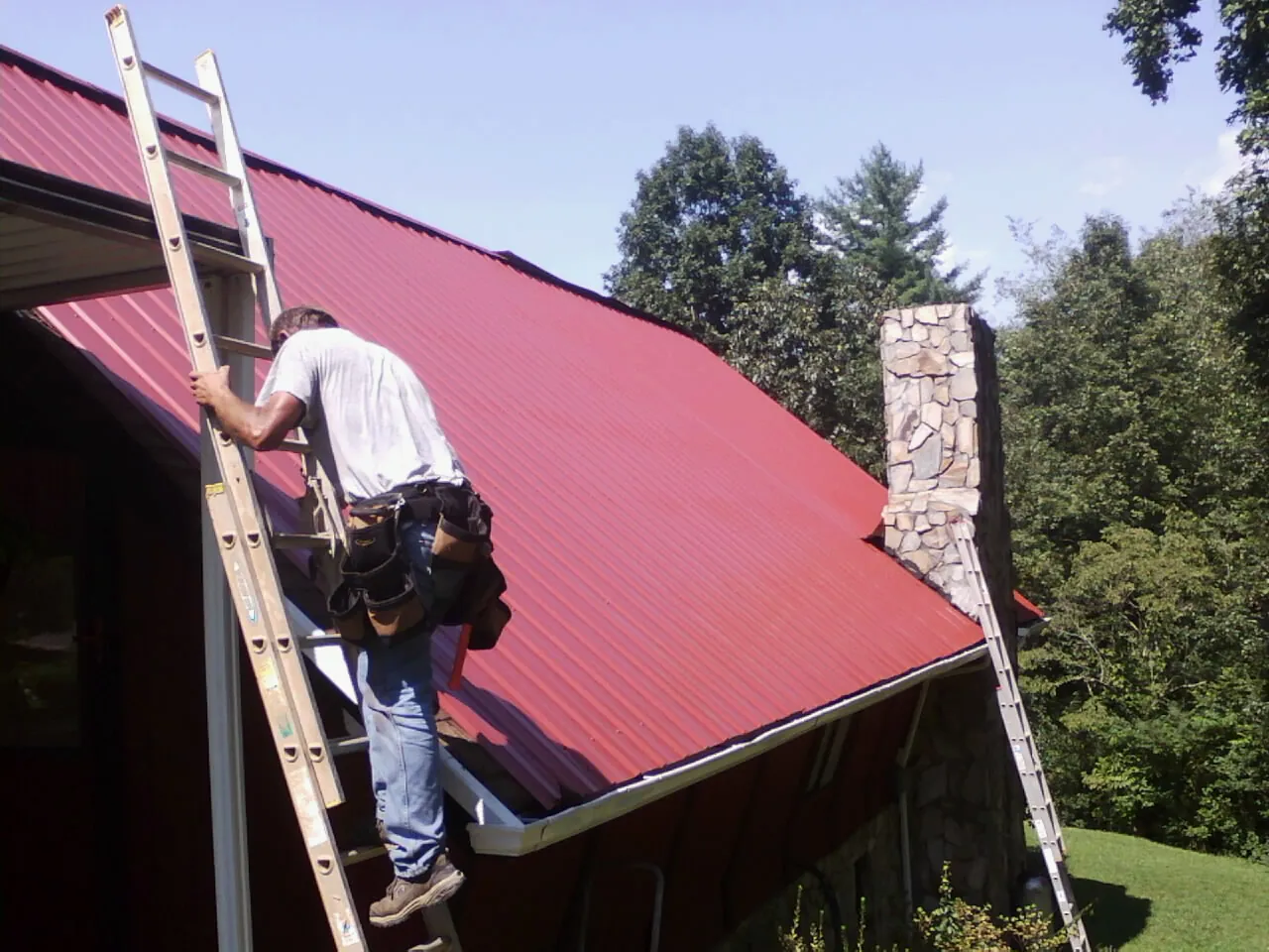 Roofer on a ladder beside a red metal roof and stone chimney — Soffit & Fascia Repair by Roofing Brothers in Rochelle